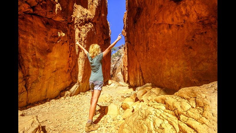 The back of a woman with long blonde hair standing with her arms raised in the middle of a gap between rock cliffs in the Australian desert
