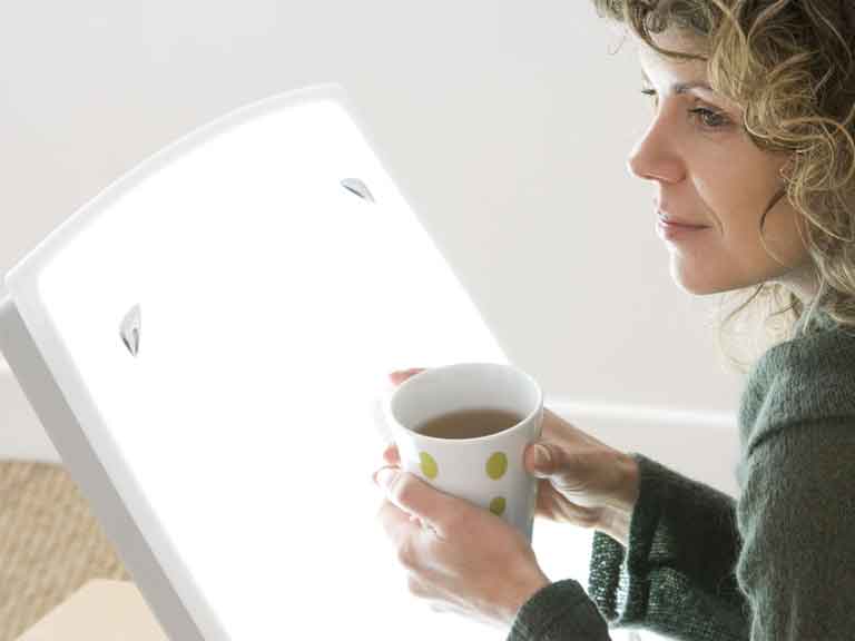 Woman sat in front of light therapy box, holding a cup