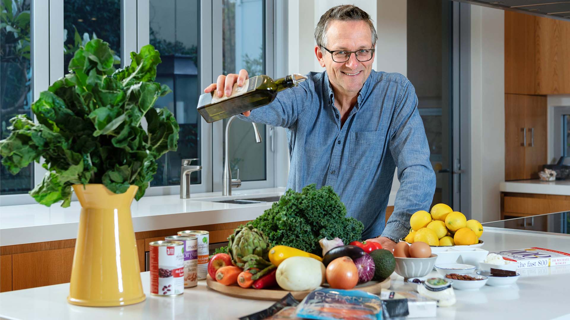 Dr Michael Mosley standing in a kitchen holding a bottle of olive oil with Mediterranean-style foods in front of him.