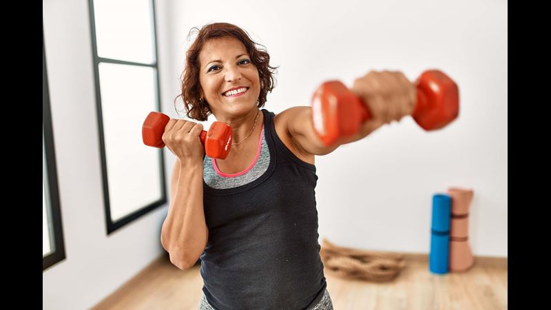 A smiling woman with a dumb bell in each hand in a gym