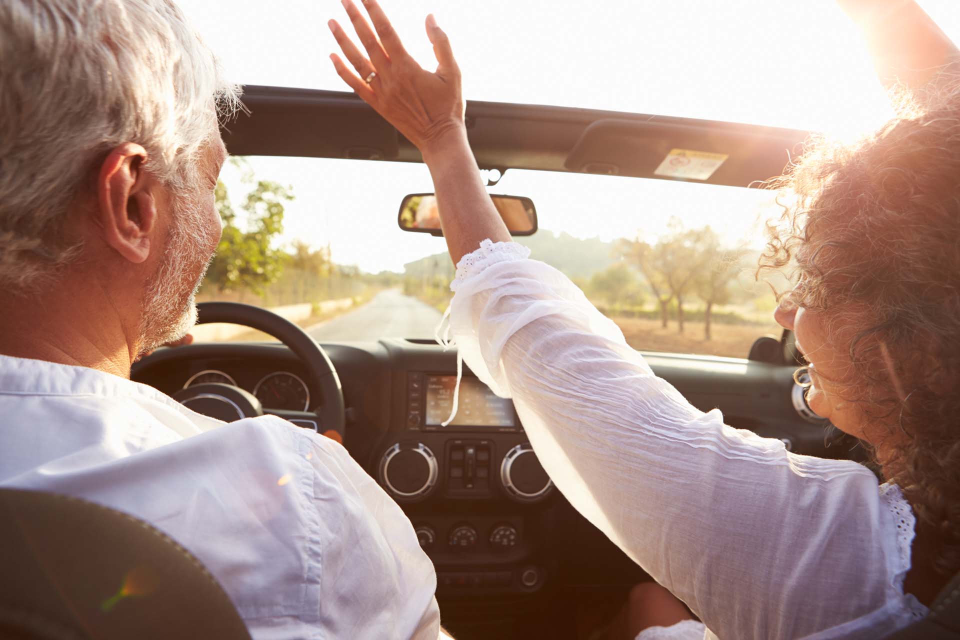 An older couple driving a convertible through sunny landscape, with the woman with her hands in the air and the man smiling