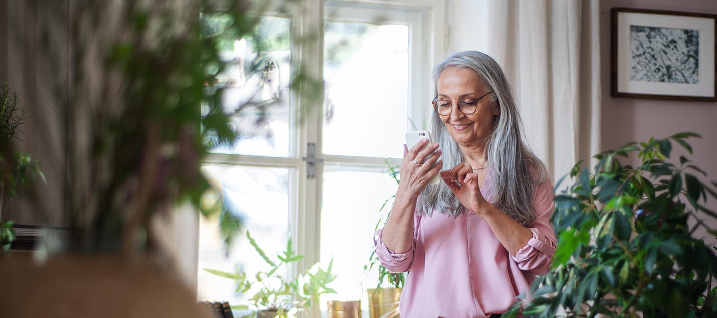Fashionable senior woman standing by desk and text messaging at home