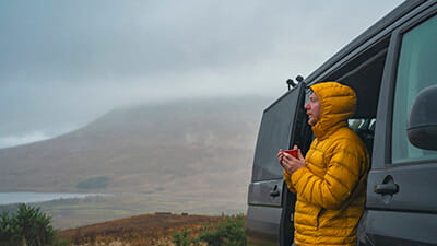 A mature man takes in the view of the Scottish Highlands on a rainy day from his campervan. He wears his hooded puffer coat and warms himself up with a cup of tea.