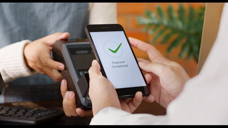 A person holding their smartphone against a contactless card reader. The screen says 'payment complete'