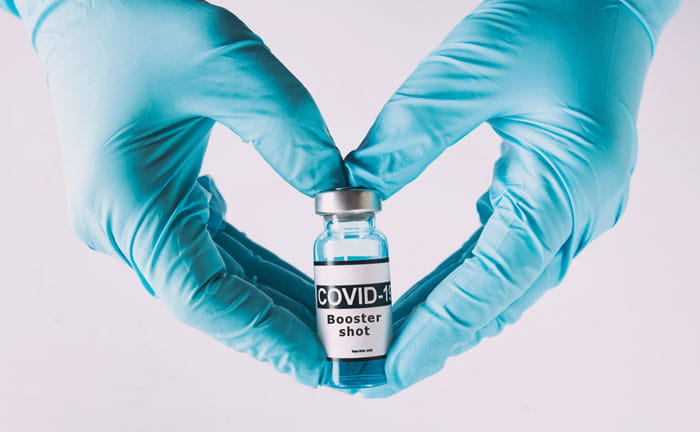Two hands positioned in a heart-shape in blue PPE gloves holding a vaccine vial