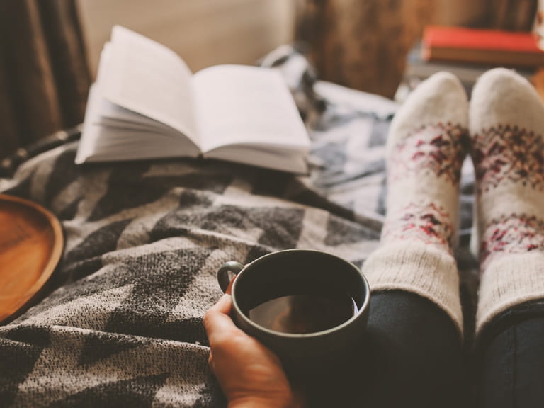 Feet in warm sock on a bed with a hand holding a mug of tea