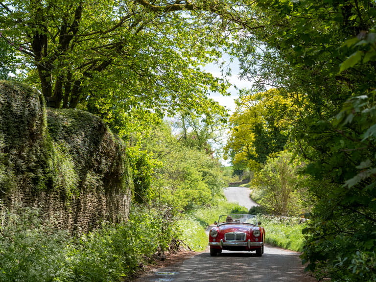 A convertible car driving along a country lane surrounded by trees