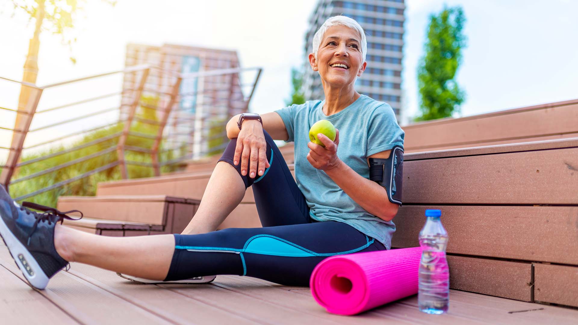 A smiling woman with cropped grey hair sitting on the ground next to a yoga mat and bottle of water, holding a green apple.