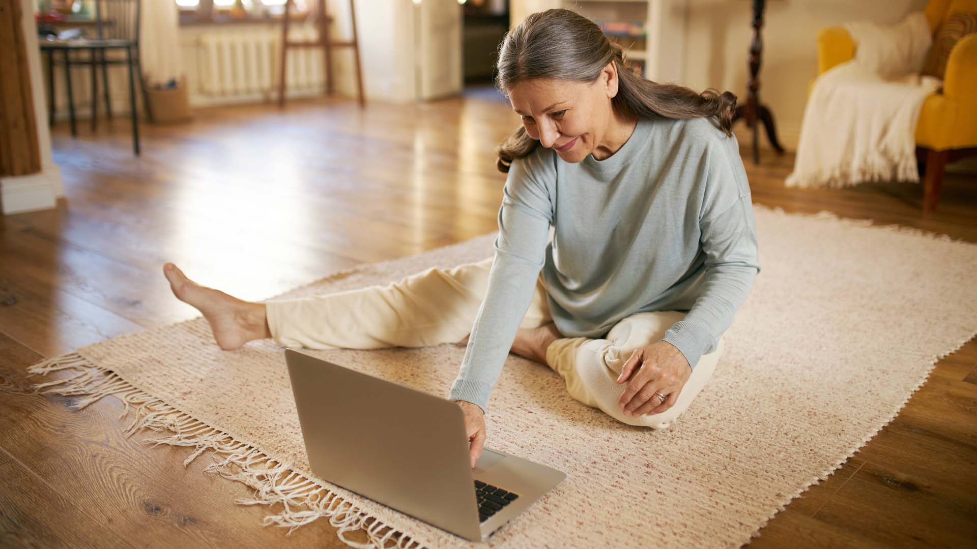 A woman sitting on a cream rug in front of a laptop in comfortable clothing
