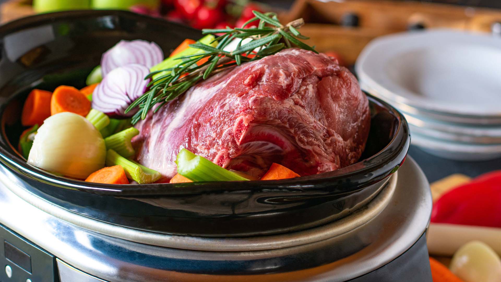 A large joint of red meat surrounded by vibrant vegetables in an oval-shaped slow cooker.