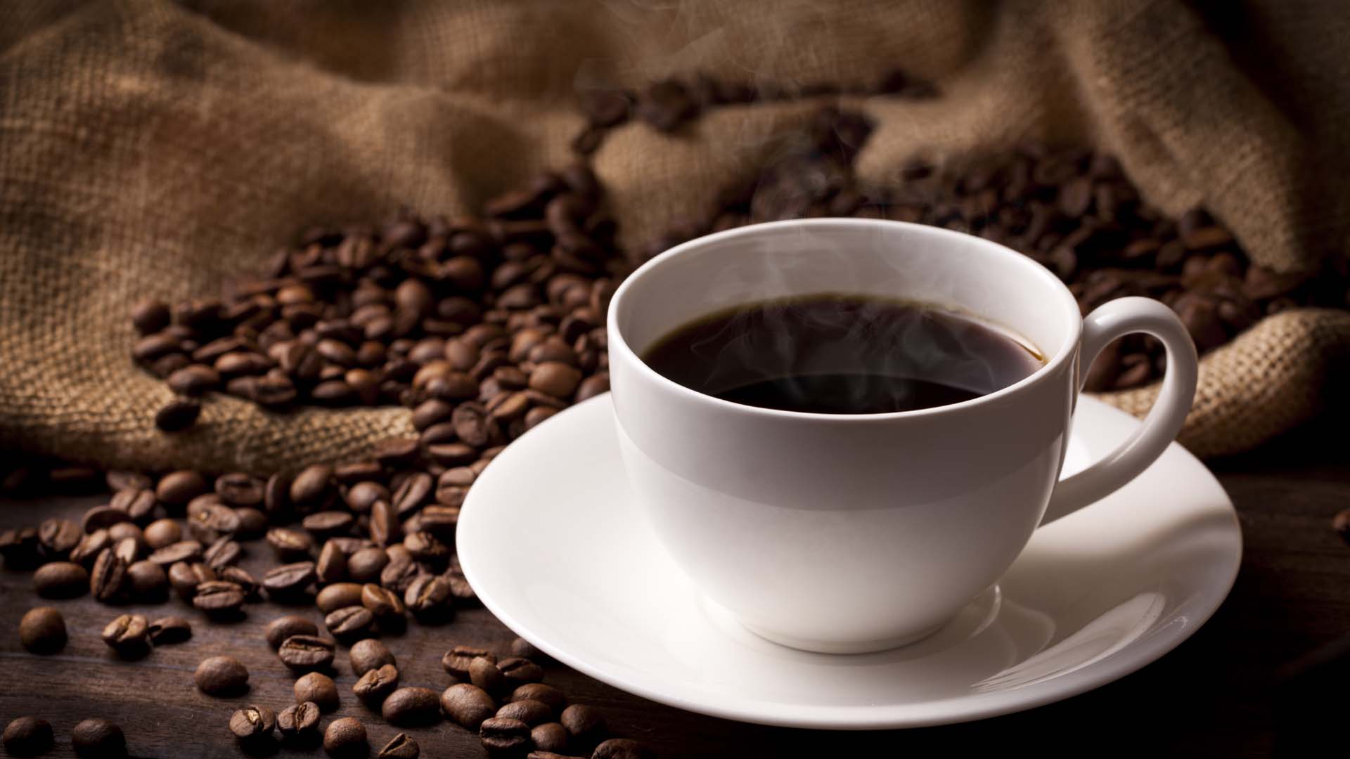 A black coffee served in a white cup and saucer, styled with a hessian sack and spilled coffee beans.