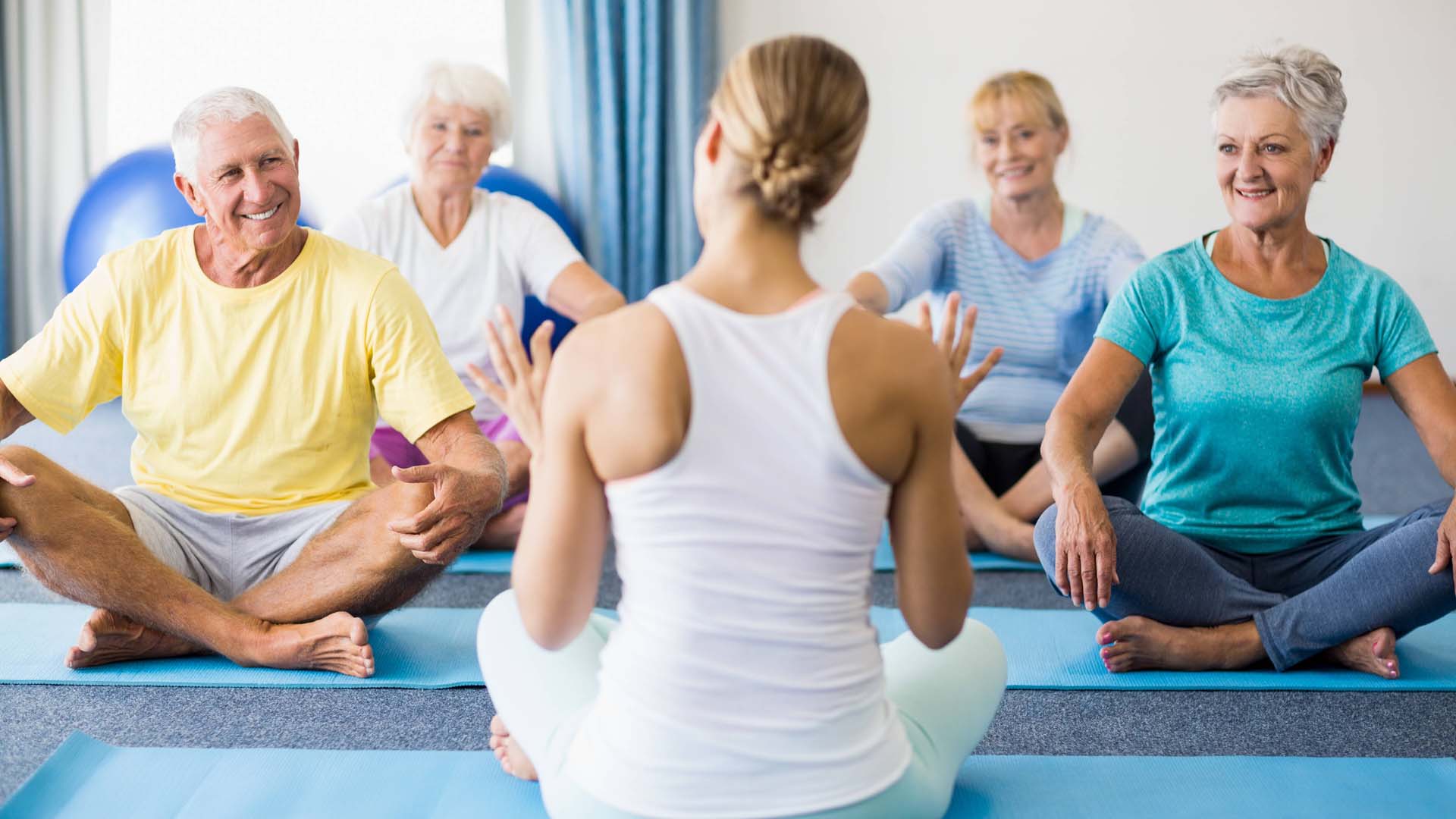 Mature yoga students sitting cross-legged on mats facing a female instructor with her back to the camera