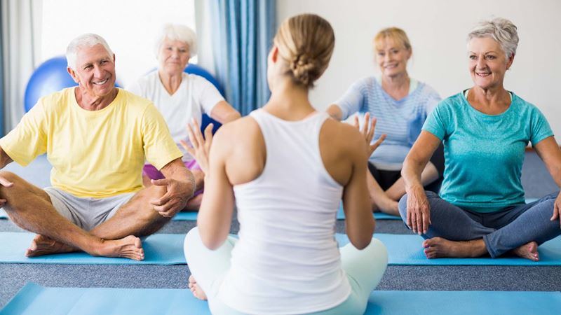 Mature yoga students sitting cross-legged on mats facing a female instructor with her back to the camera