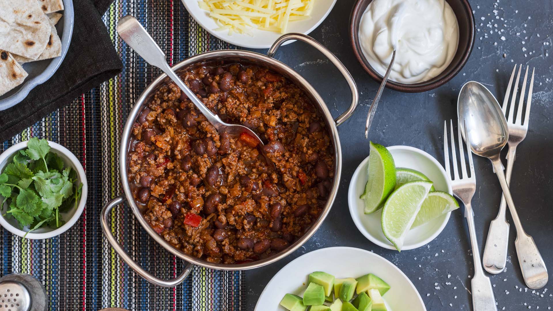 Flat lay image of a large silver dish of chilli con carne surrounded by smaller plates of lime, yoghurt, naan breads and coriander, with silver cutlery.
