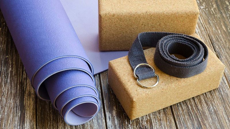 A blue rolled-up yoga mat, two brown yoga blocks and black strap displayed on a wooden floor
