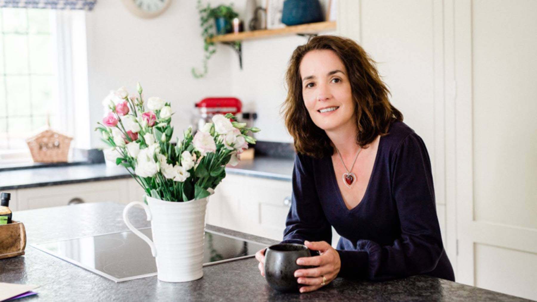 A woman leaning on a kitchen island holding a mug with a vase of flowers next to her