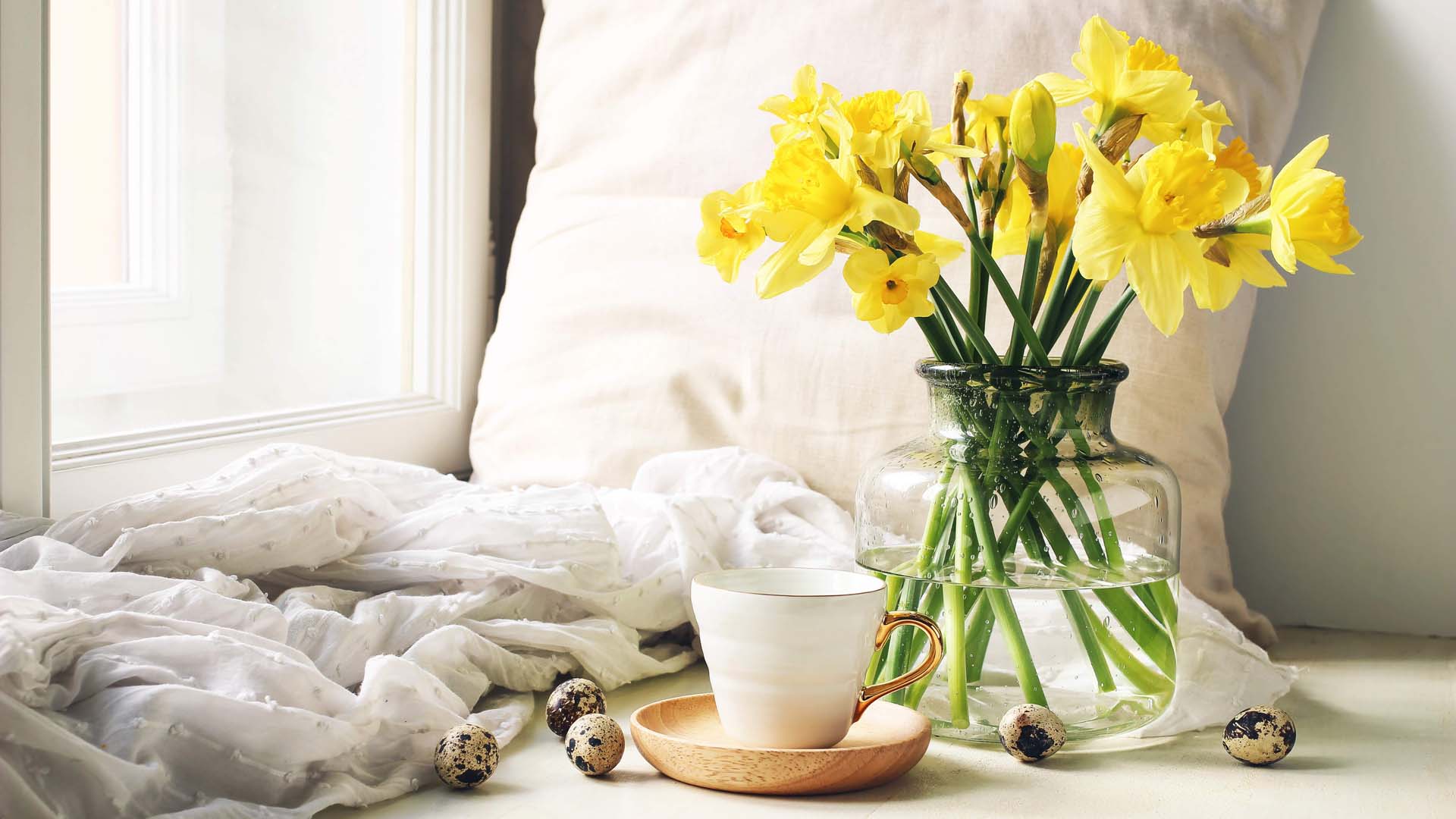 Cup of coffee, wooden plate, quail eggs and vase of daffodils on windowsill.
