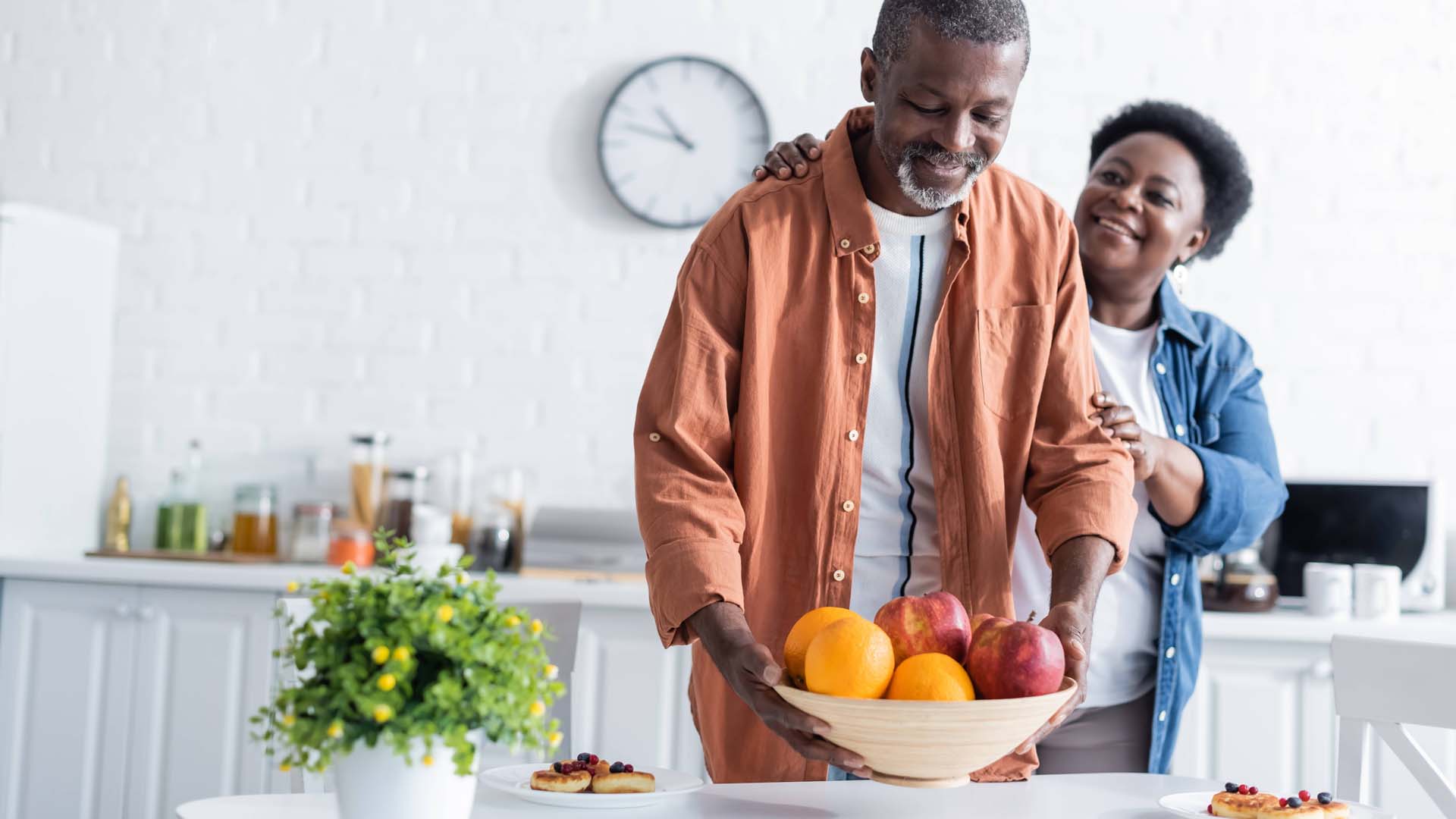 Man putting bowl with fruits on breakfast table near smiling wife