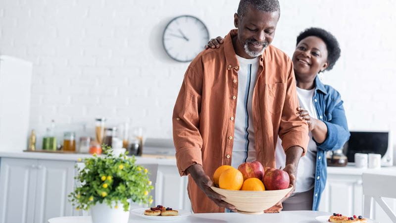 Man putting bowl with fruits on breakfast table near smiling wife