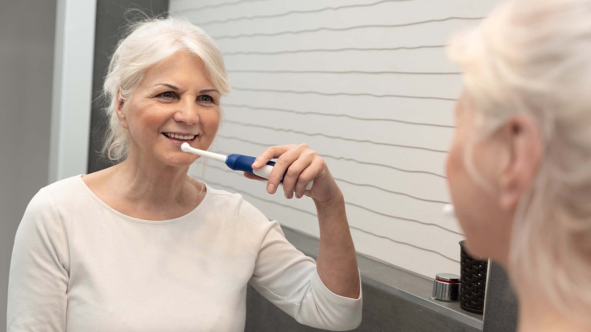 An older white haired woman smiles while brushing her teeth