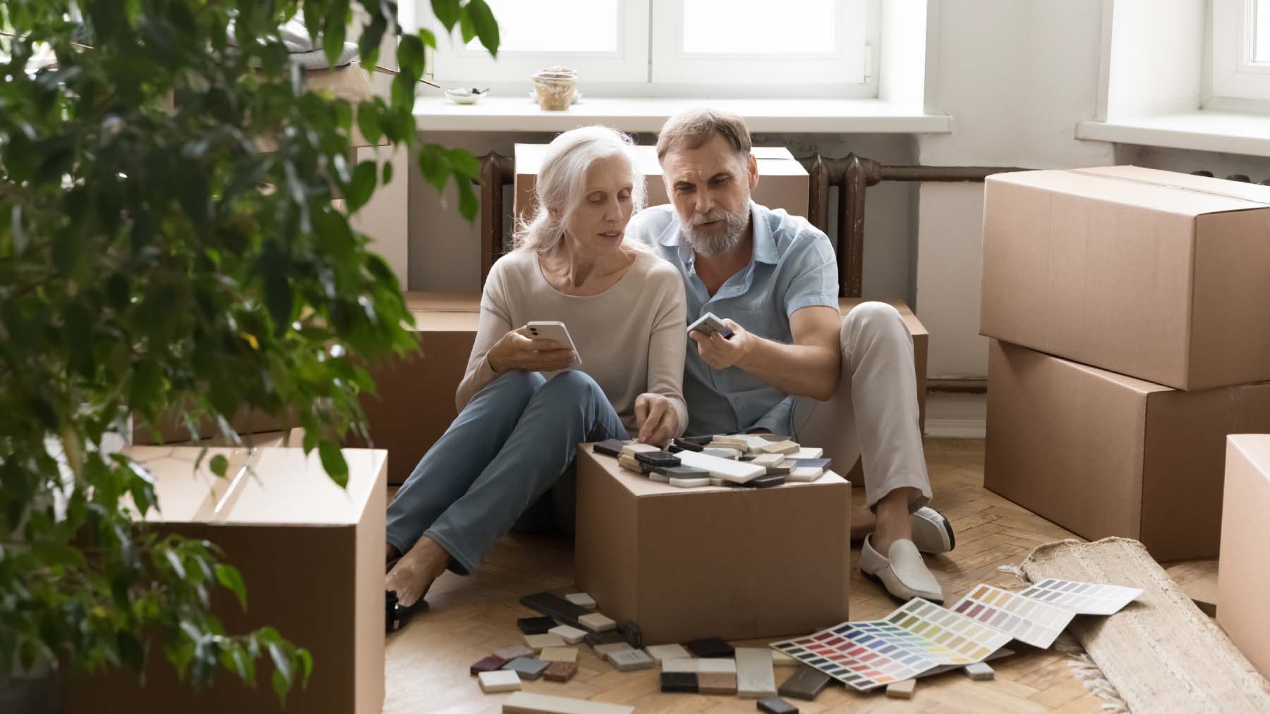 A couple sit on the floor indoors, surrounded by moving boxes and paint swatches