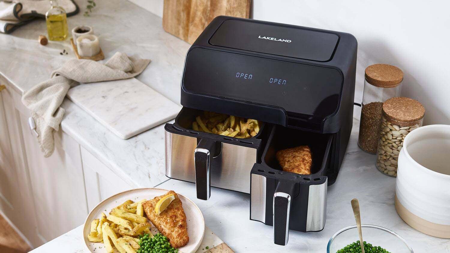 The Lakeland Dual Basket Air Fryer on a white worktop next to a plate of fish and chips and other cookware.
