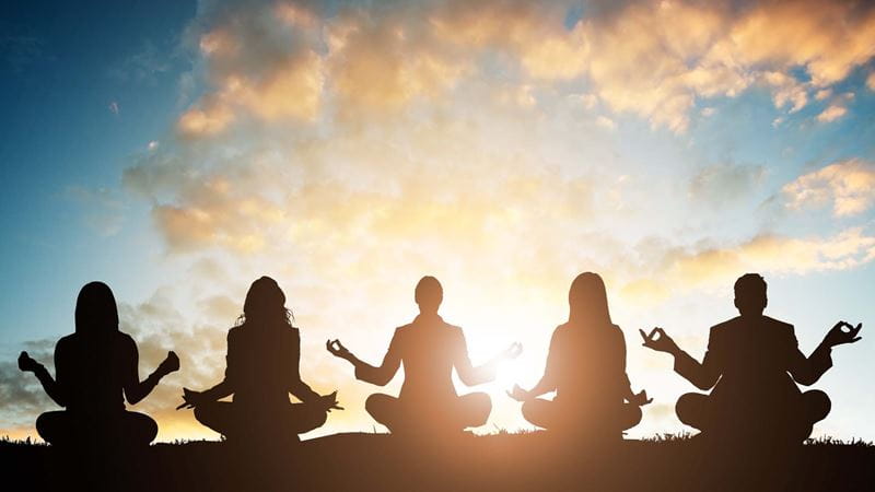 Silhouette of five people sitting cross-legged practicing yoga against a blue sky and sunset