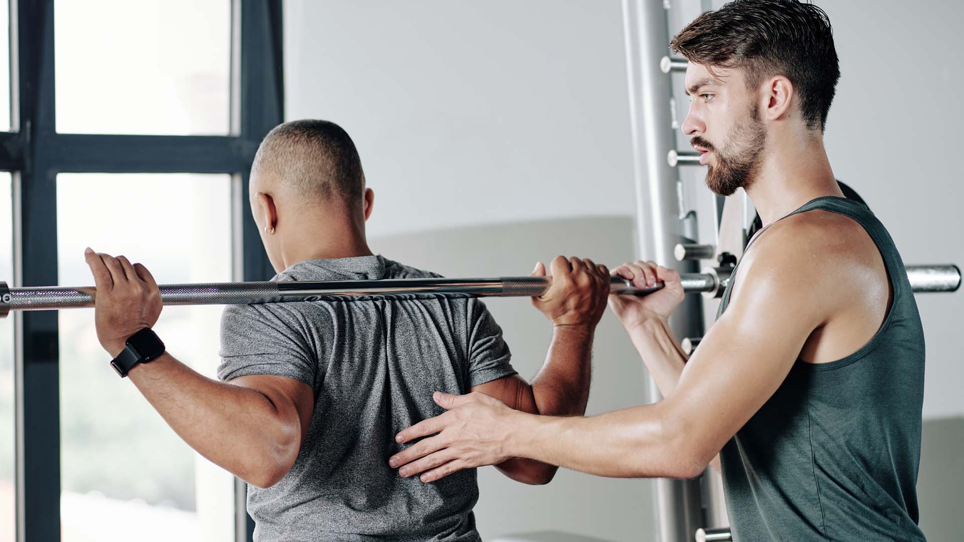 A man lifts a bar of weights in a gym environment with the help of a young male personal trainer