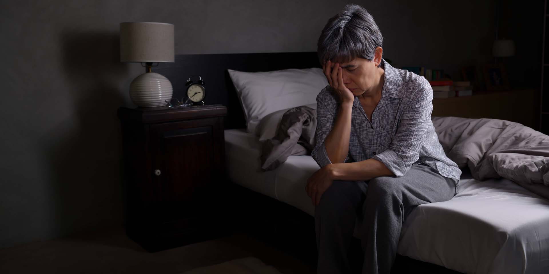An older woman sitting on the edge of an unmade bed with head in hands as if struggling to sleep