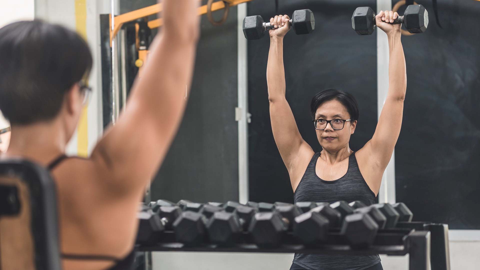 A woman looking in a gym mirror while doing a shoulder press with two dumbbells