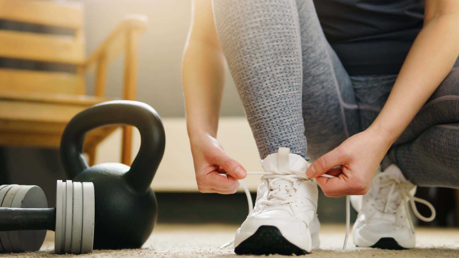 Close-up of a woman in grey leggings tying her trainers next to a kettlebell and weight 