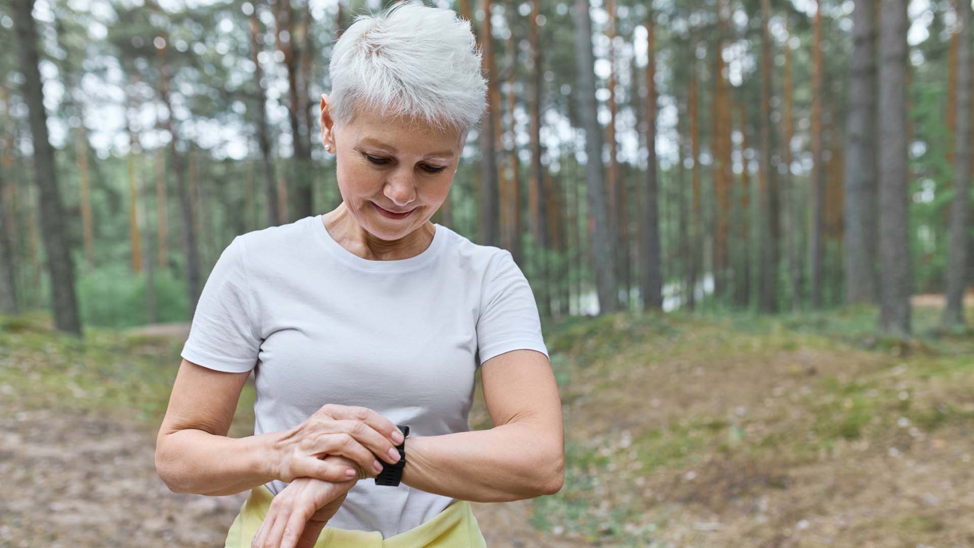 A woman with cropped white hair checks a fitness watch while exercising in a woodland area