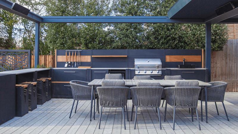 A grey table with 8 chairs in the centre of L shaped outdoor kitchen unit in charcoal with wooden accents, covered by a retractable roof