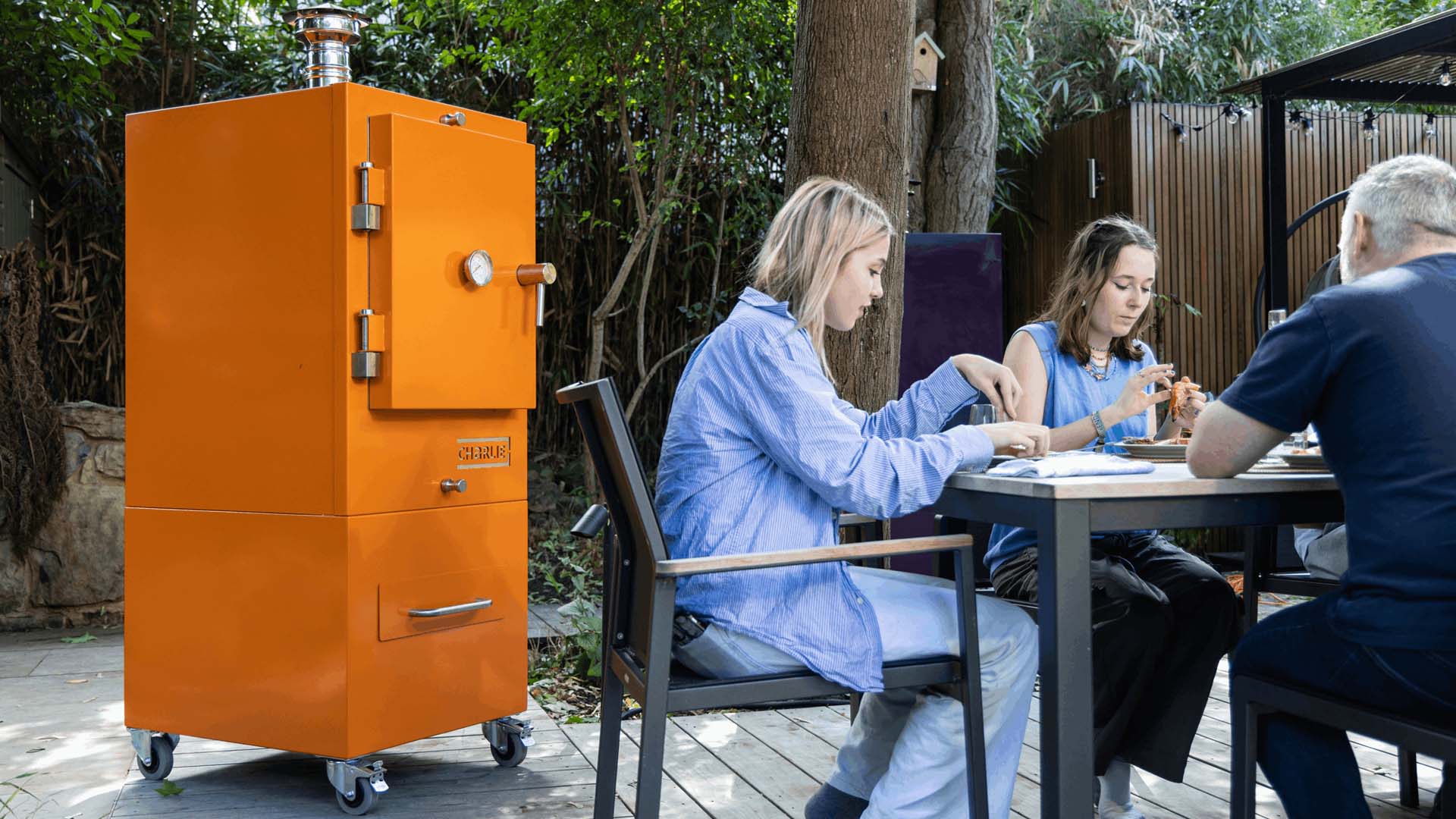 An orange, vertical cooker on wheels next to 3 people dining at an outdoor table