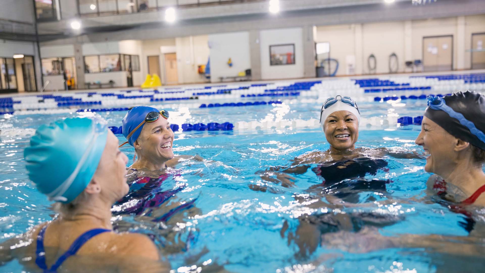 A group of women wearing swimming caps and goggles chatting in a swimming pool