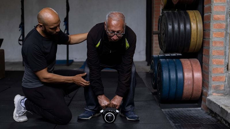 An older person is assisted by their personal trainer as they lift weights