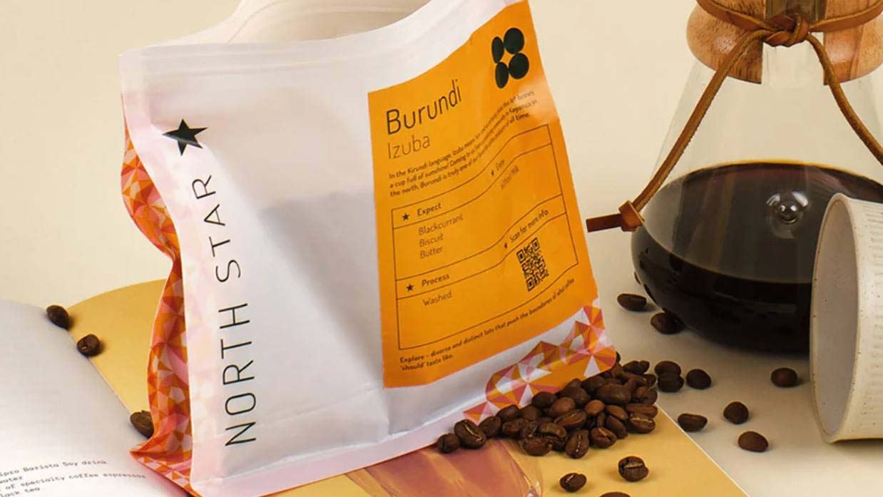 Close-up of a bag of North Star coffee beans displayed on an open book, with a coffee pot brewing in the background and a scattering of coffee beans.