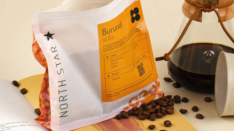 Close-up of a bag of North Star coffee beans displayed on an open book, with a coffee pot brewing in the background and a scattering of coffee beans.