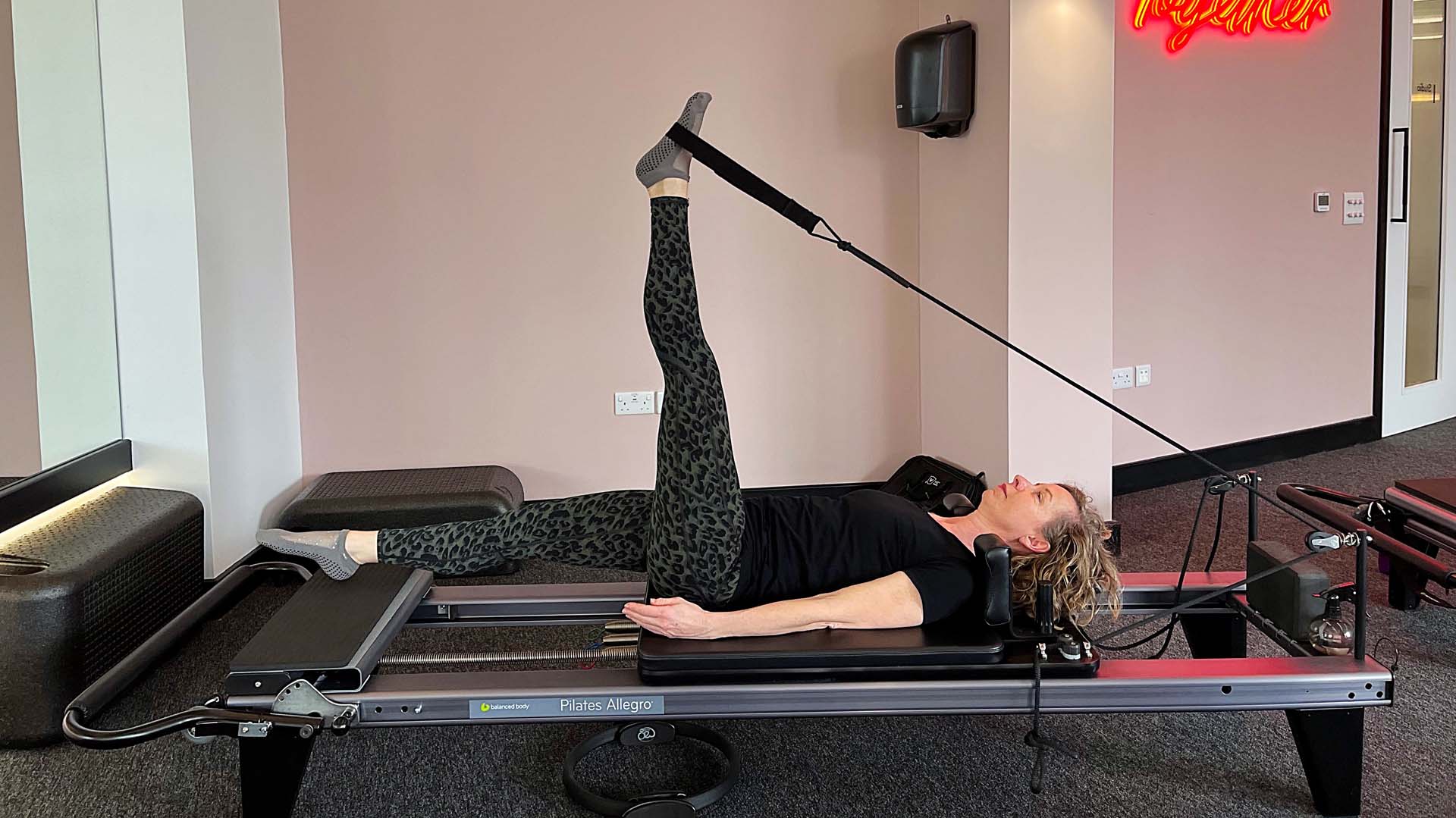A woman in sports leggings lying on a reformer machine using an elastic band to stretch a leg in the air.