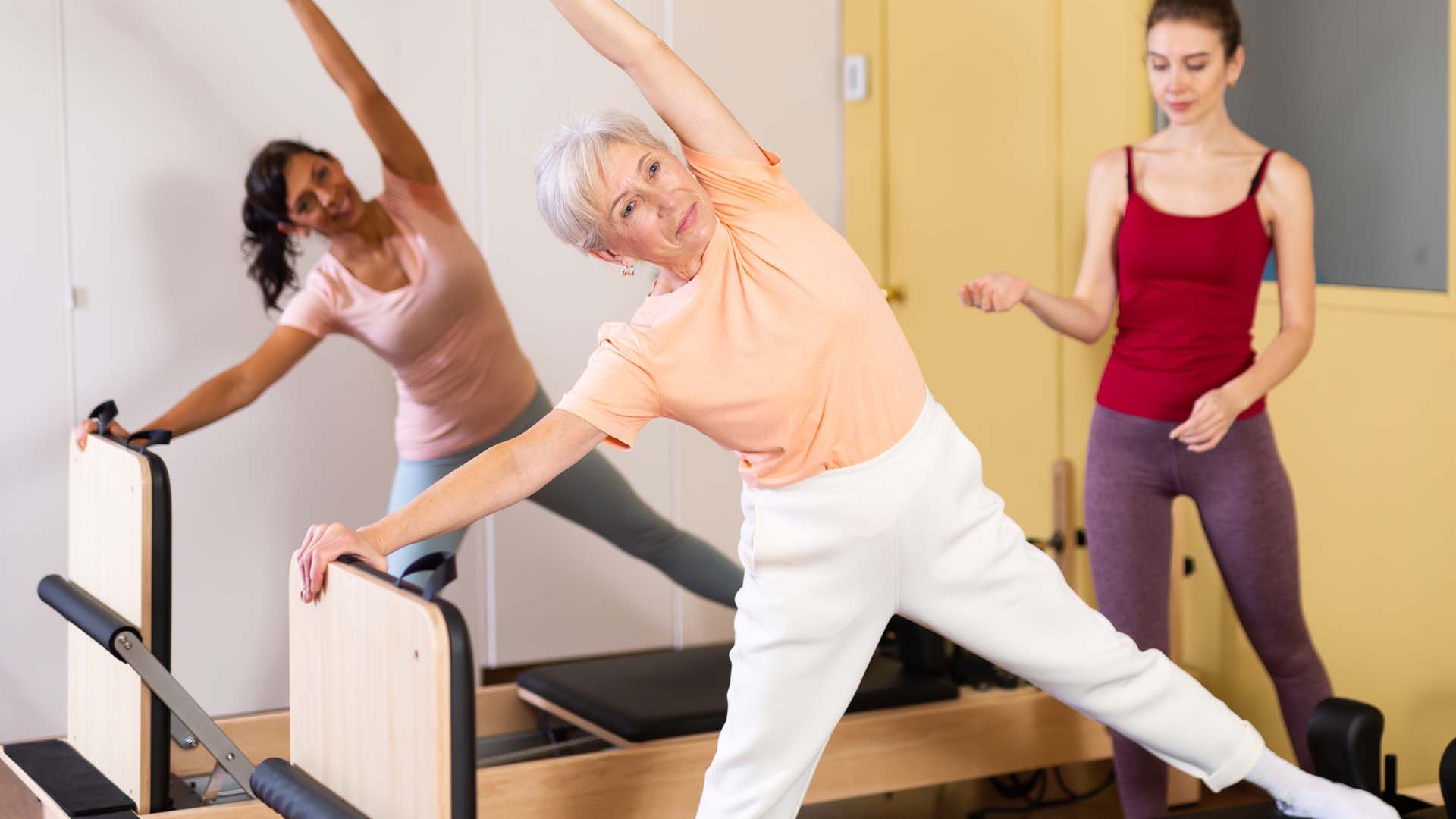 Two older women practicing reformer Pilates by standing on reformer machines and stretching while an instructor oversees.
