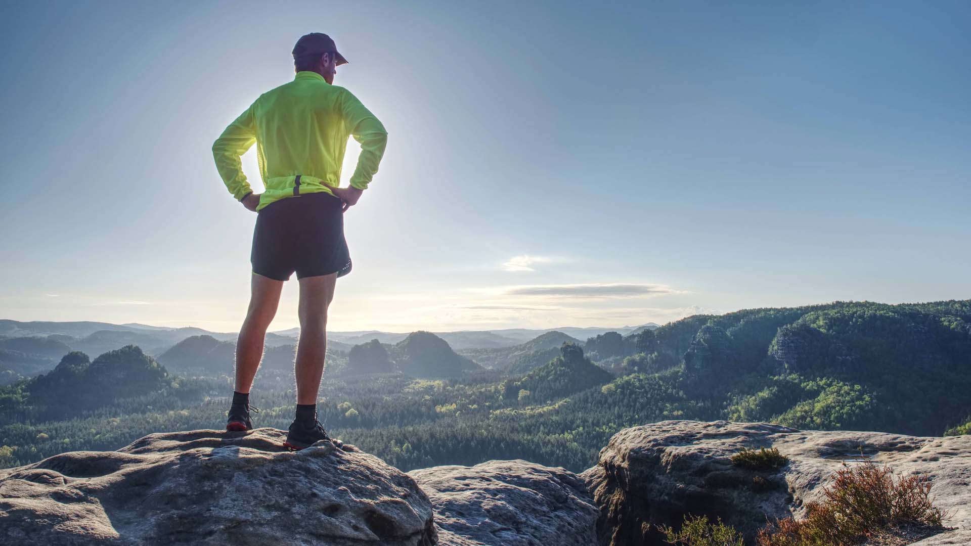 A man wearing a bright exercise top stands on top of a rock to admire a sweeping countryside vista on a sunny day