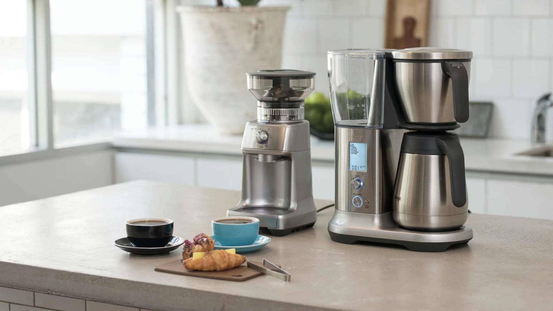 A drop coffee machine and coffee grinder on a kitchen island counter, along with two mugs of coffee and a croissant