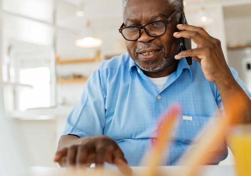 An old black man is sitting at his home office, talking on the phone with a customer support worker and using his laptop