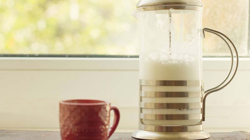 A French press containing frothed milk standing in front of a window beside a brown mug.