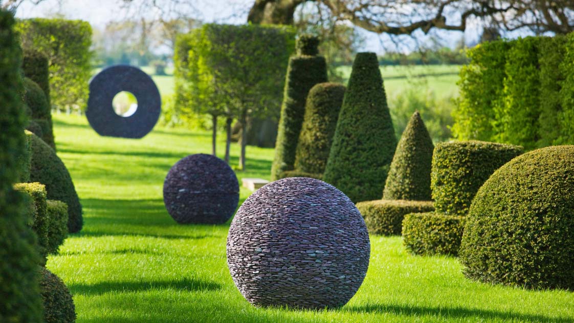 A large garden lawn lined with topiary hedges and decorated with large spherical and circular sculptures on a sunny day.