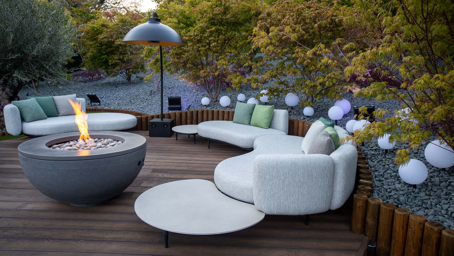 A curved garden deck arranged with curved grey sofas and a fire pit with trees in the background.