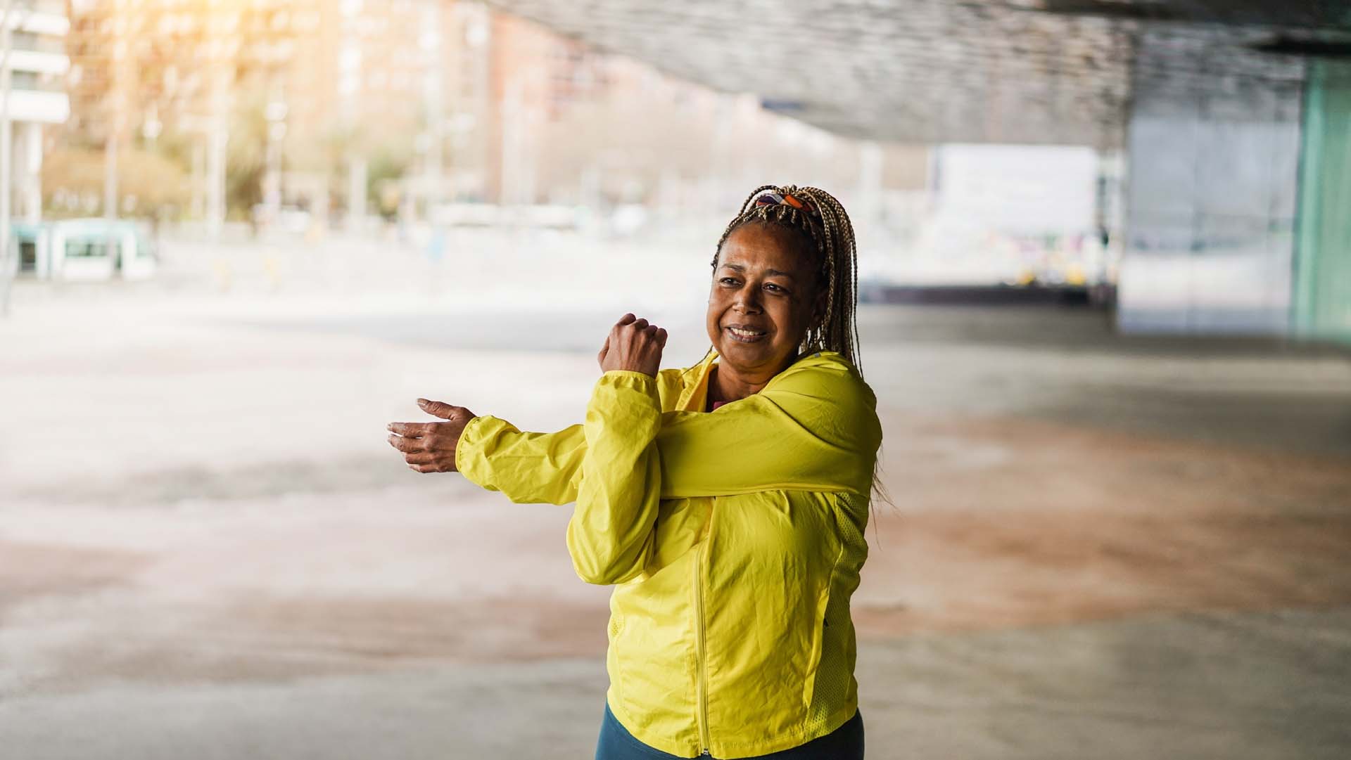 A woman in a yellow sports top stretches an arm across her chest while exercising in an urban area