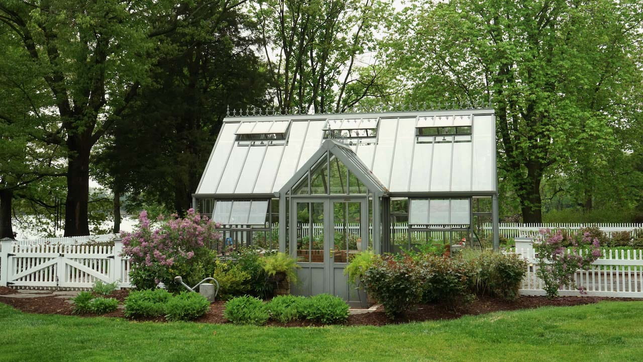 Glass greenhouse in the middle of a white-fenced area surrounded by large trees, grass field and plant beds
