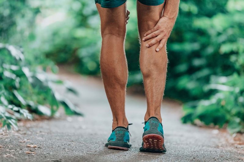 Lower half of a man who is holding his calf as if in pain while exercising outdoors on a path surrounded by greenery