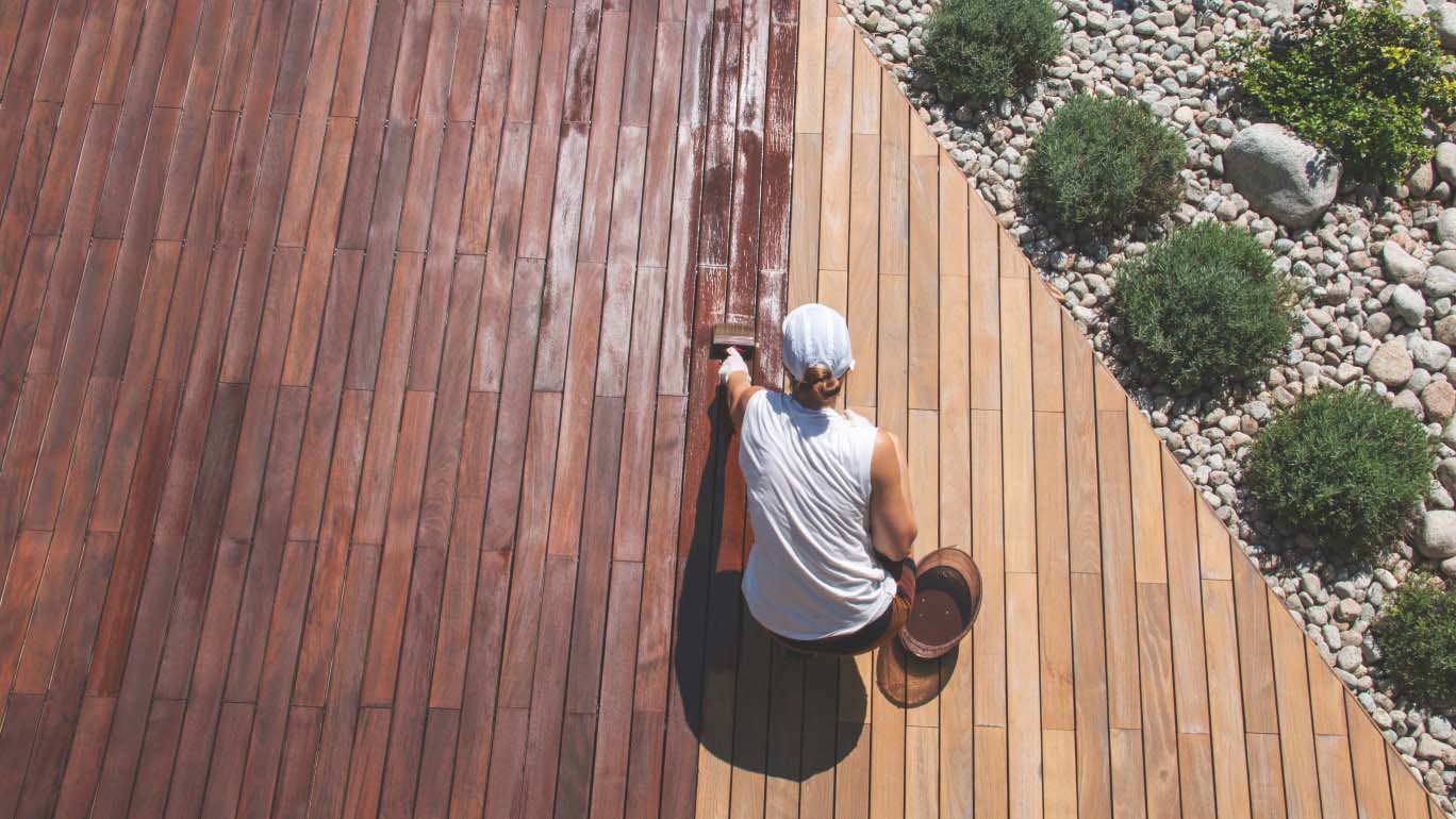 Someone varnishing a wood deck in the sun bordering a gravel garden